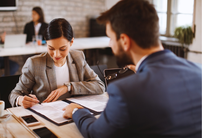 A woman in a gray blazer signing documents while sitting at a table with a man in a blue suit. They appear to be engaged in a discussion in a modern office setting.