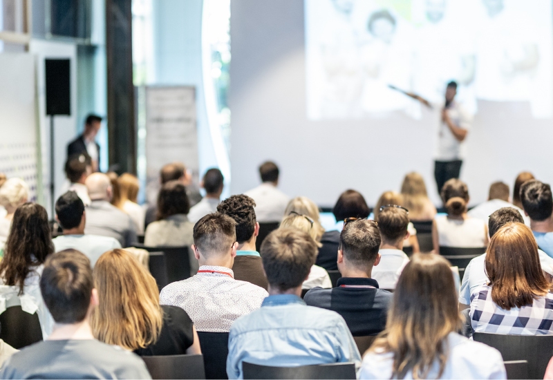 An audience of professionals seated in a conference hall, facing a speaker presenting a slide show on a large screen. Attendees engaged in the presentation, some taking notes.