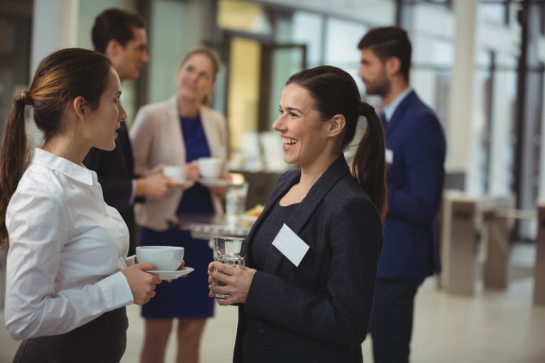 Two women engaged in conversation at a networking event, one holding a cup and the other with a drink. In the background, other attendees are chatting and a table with refreshments is visible.