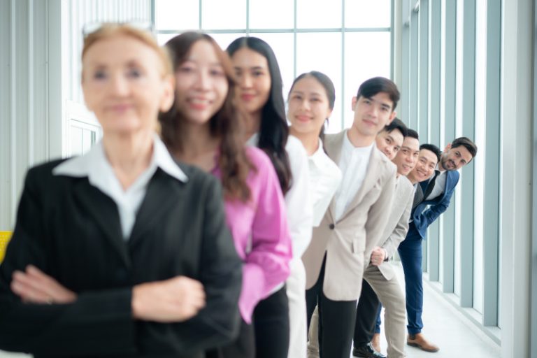 A diverse group of professionals standing in a line in a modern office space, with a large window in the background. The individuals are dressed in business attire, showcasing various styles. The focus is on the individuals, while the foreground is slightly blurred.